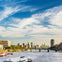 A cityscape with a river, several boats, a bridge, and buildings in the background under a partly cloudy sky.