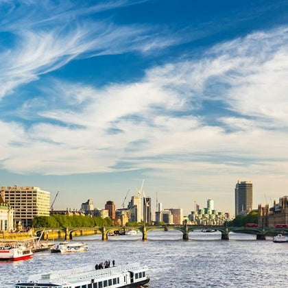A cityscape with a river, several boats, a bridge, and buildings in the background under a partly cloudy sky.