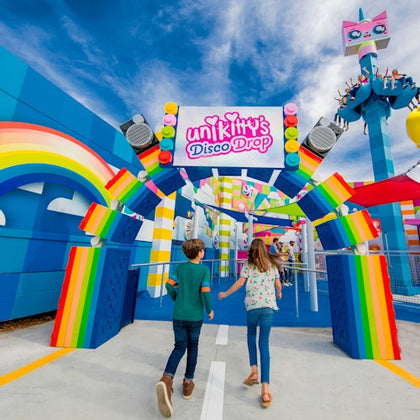 Two children walk towards a colorful amusement park ride entrance with rainbow decorations and a sign reading "Unikitty's Disco Drop."