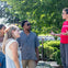 A group of people outdoors, standing and talking near greenery under sunlight, with one person gesturing as they speak.