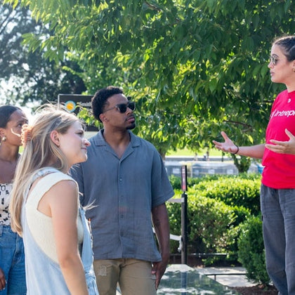 A group of people outdoors, standing and talking near greenery under sunlight, with one person gesturing as they speak.