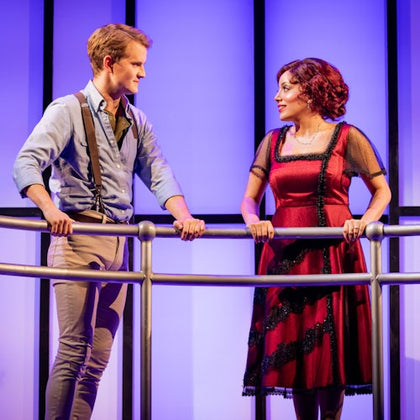 Two actors stand on a balcony set against a blue-lit background, engaging in conversation.