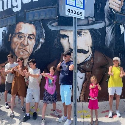 A group of people in front of a mural depicting two men with intense expressions. They are posing playfully and pointing at the camera.