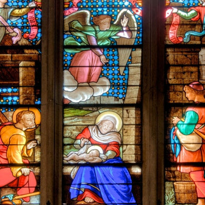 A stained glass window depicting a nativity scene with Mary, Joseph, baby Jesus, an angel, and shepherds in vibrant colors.