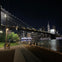 Night view of a lit-up bridge over a river with a city skyline in the background and people walking along the promenade.