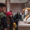 Two individuals observing a sculpted bust with eclectic adornments in a museum setting, with other visitors in the background.