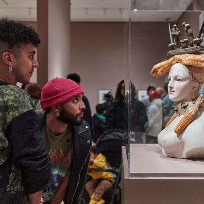 Two individuals observing a sculpted bust with eclectic adornments in a museum setting, with other visitors in the background.