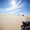 People riding ATVs on sandy dunes under a bright sun with clear skies; tire tracks visible in the sand.