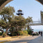 People walking under an arched passage with a lighthouse, trees, and a bridge visible in the background.