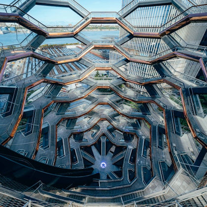 A multi-level hexagonal structure with interconnected stairways, surrounded by modern buildings and overlooking a waterfront.
