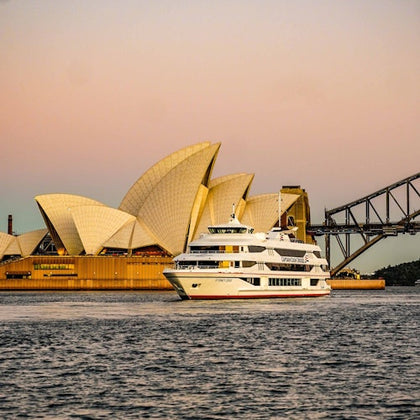 A ferry on the water near the Sydney Opera House and Sydney Harbour Bridge during sunset, with a pastel sky.