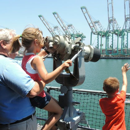 A girl looks through a telescope on a waterfront observation deck, assisted by a man, with a boy nearby waving.