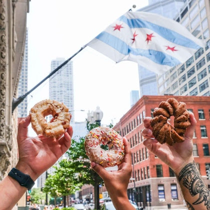 Three hands holding differently decorated donuts with the Chicago flag and city buildings in the background.
