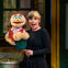 A performer holding a puppet with brown hair and a cheerful expression on a stage with a set resembling an urban environment.