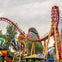 A vibrant amusement park scene with a red roller coaster, colorful hot air balloon ride, trees, and a cloudy sky.