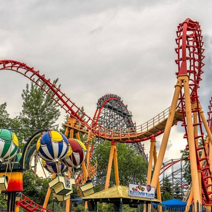 A vibrant amusement park scene with a red roller coaster, colorful hot air balloon ride, trees, and a cloudy sky.