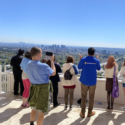People stand on a viewing platform, some taking photos, overlooking a cityscape with clear skies.