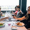 Two people clinking wine glasses at a dining table in a restaurant, with others dining in the background.