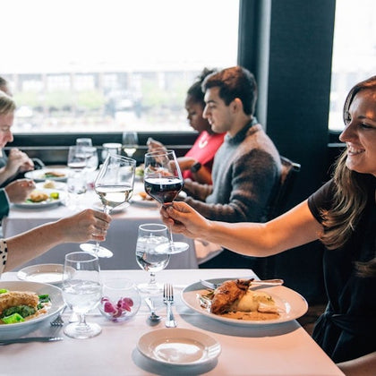 Two people clinking wine glasses at a dining table in a restaurant, with others dining in the background.