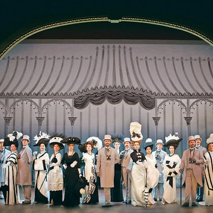 A group of people dressed in formal, period-style clothing on stage with a decorative backdrop.
