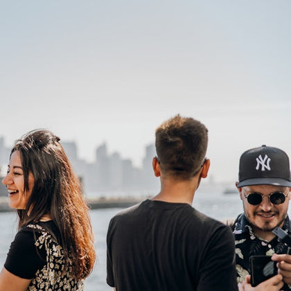 Three people standing outdoors with a city skyline in the background. One is smiling, another facing away, and one holding a phone.