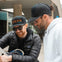 Two men in sunglasses and caps, one in a black jacket and the other in a white hoodie, look at a paper in an urban setting.