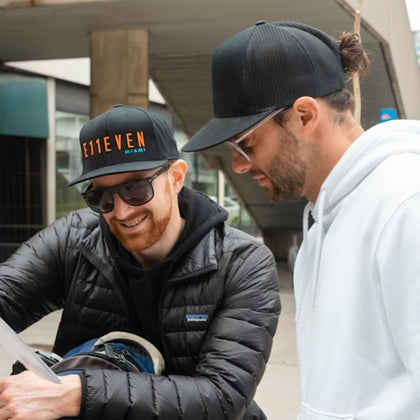 Two men in sunglasses and caps, one in a black jacket and the other in a white hoodie, look at a paper in an urban setting.