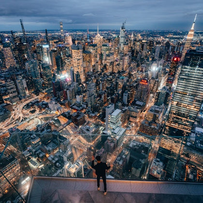 A person stands on a high observation deck overlooking a cityscape of lit skyscrapers and streets at dusk.
