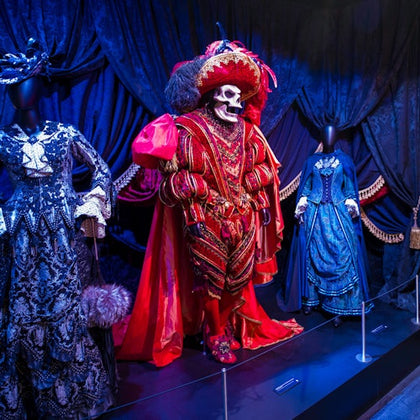 Three elaborate costumes on mannequins: a red skeleton in the center, flanked by two Victorian-style dresses, against a dark, draped background.