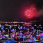 A nighttime scene of a harbor filled with boats lit by colorful lights, with fireworks visible in the sky above.