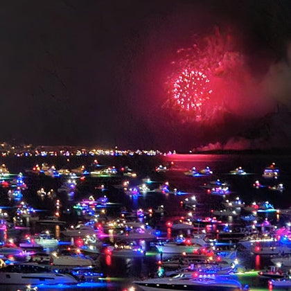 A nighttime scene of a harbor filled with boats lit by colorful lights, with fireworks visible in the sky above.