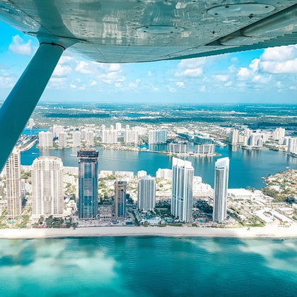 View from an airplane over a coastal city with tall buildings, water bodies, and a beach under a partly cloudy sky.