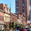 A city street with historic brick buildings, trees, hanging lights, a pedestrian, and modern high-rise buildings in the background.
