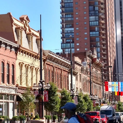 A city street with historic brick buildings, trees, hanging lights, a pedestrian, and modern high-rise buildings in the background.