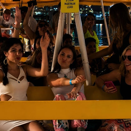 A group of people enjoying a nighttime boat ride, with some raising their hands and smiling, under yellow lighting.