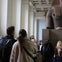 Visitors in a museum observe and photograph a large stone sculpture displayed in a spacious gallery.