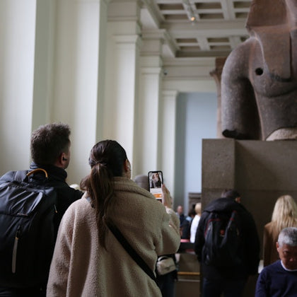 Visitors in a museum observe and photograph a large stone sculpture displayed in a spacious gallery.