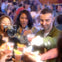 People raising glasses for a toast in a crowded, lively setting. One person is using a film camera to capture the moment.