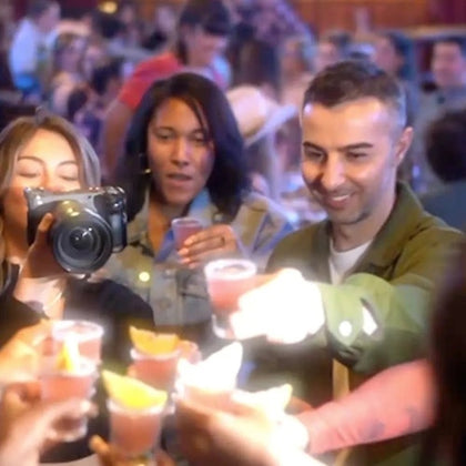 People raising glasses for a toast in a crowded, lively setting. One person is using a film camera to capture the moment.