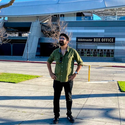 A man in a green shirt stands on a sidewalk near a Ticketmaster box office, with a modern building and leafless trees in the background.