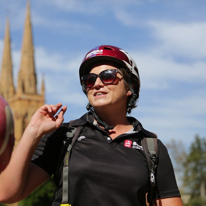 A person wearing sunglasses and a helmet stands outdoors, with a church spire in the background on a sunny day.