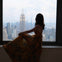 A person in a floral dress sits on a windowsill overlooking a cityscape with the Empire State Building visible.