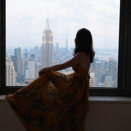 A person in a floral dress sits on a windowsill overlooking a cityscape with the Empire State Building visible.