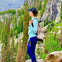 A person wearing a light blue top and navy pants with a backpack stands on a rock in a forested mountainous landscape.