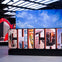 Large "CHICAGO" sign with letters displaying city landmarks, food, and buildings, backed by a screen showing a blue sky with clouds.