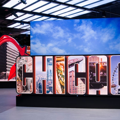 Large "CHICAGO" sign with letters displaying city landmarks, food, and buildings, backed by a screen showing a blue sky with clouds.