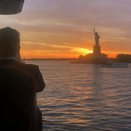 A person photographs the Statue of Liberty at sunset, with water and a boat visible in the foreground.