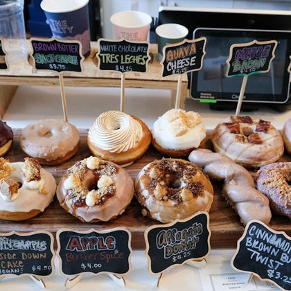 An assortment of gourmet donuts displayed with labels indicating different flavors such as brown butter, white chocolate tres leches, and maple bacon.