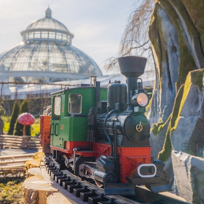 A miniature green and red locomotive on a track in a garden setting with a large glass conservatory in the background.