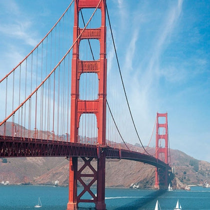 A red suspension bridge spanning a body of water, with sailboats and hills in the background under a blue sky.